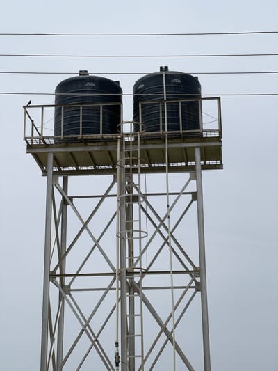 Two large, black cylindrical water tanks are elevated on a metal scaffolding structure against a pale, overcast sky. The tanks are stacked vertically, with the lower tank positioned slightly forward of the upper tank. Each tank has a textured surface and appears to be made of plastic. Metal railings and a ladder are attached to the scaffolding around the tanks, suggesting they are for public access or maintenance.
Thin, dark wires run vertically across the left side of the image, creating a pattern against the sky. The metal structure is constructed from interconnected beams and supports, forming a geometric lattice. Some areas of the metal show signs of rust.
There is no discernible activity taking place. The weather appears to be overcast or foggy, providing diffused light and a muted color palette. The scene is likely an outdoor setting, possibly in a rural or industrial area, given the elevated tanks and scaffolding.
Visible text on the upper tank, though partially obscured, appears to be a label or inscription. The text is white against the black surface. The overall mood is utilitarian and functional. The image's orientation is rotated 90 degrees counter-clockwise.