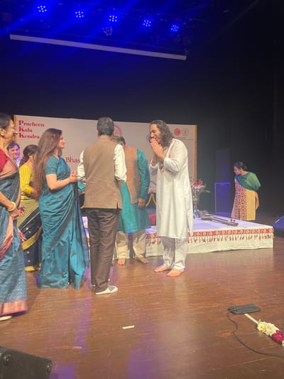 The image shows a group of people on a stage in the Pracheen Kala Kendra in Chandigarh, India (latitude: 30.733, longitude: 76.79). There are five people on the stage: a man in a white shirt with his hands clasped in prayer, a woman in a blue sari, and three men behind them. The man in the white shirt has long hair and is barefoot. The woman is looking at the man in the white shirt. The three men are all wearing casual clothing. The man in the brown jacket is looking down at the woman in the blue sari. The man to his right is looking at the man in the white shirt. There is a man in a teal shirt in the background on the stage. There is a sign on the wall behind the people that reads "Pracheen Kala Kendra" and "Bhas" in Hindi. There is a musical instrument on the stage. There are lights overhead, and the stage is made of wood. There is a woman in a colorful dress in the background who is looking down at the stage. There is a black speaker on the stage and a cord running along the floor to the lower left-hand side of the image. There is a white flower garland on the stage.