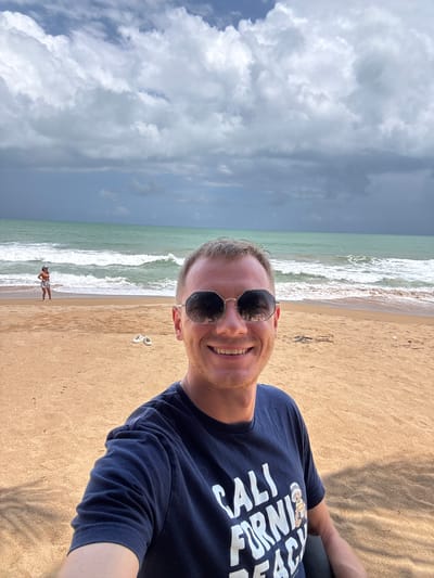 A selfie taken on a beach shows a smiling man with short brown hair wearing sunglasses and a navy blue t-shirt with white lettering that reads "CALI FORNI BEACH" and a small cartoon bear. He is positioned in the foreground. Behind him, the beach is a light tan color. Further back, a woman in an orange top and white shorts stands near the water's edge, looking out at the sea. White waves are breaking on the shore. The ocean is a greenish-blue. The sky is filled with dramatic, dark gray and white clouds. In the middle ground on the sand, a pair of white flip-flops are visible. The scene is likely in the city of Ban Khuk Khak, Thailand, given the tropical beach environment.