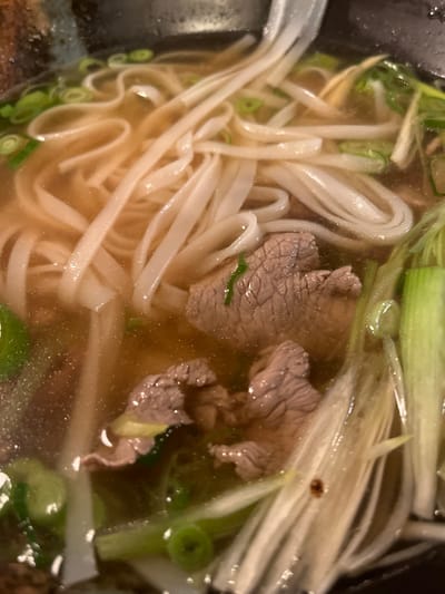 A close-up shot captures a bowl of Vietnamese Pho. Flat rice noodles intertwine with thin slices of beef in a clear, light broth. Chopped green onions are scattered throughout, adding a fresh element. The image focuses on the textures and colors within the bowl, highlighting the noodles' creamy white hues against the broth's golden transparency. The beef appears partially cooked, revealing its grain.