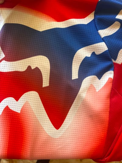 The image shows a close-up of a fabric with a red, white, and blue graphic design that appears to be the Fox Racing logo. The fabric has a textured, perforated surface. There is a seam visible on the right side of the image, where a solid red fabric is attached. The lighting is bright, creating highlights and shadows on the fabric's texture. The scene is indoors, with a blurry background that suggests a domestic setting, possibly a folded garment on a surface. The image does not depict any specific location in the city of Vasilyevo, Russia.
