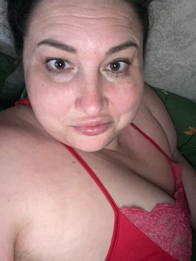 The image is a close-up, horizontally oriented, selfie-style photo of a person lying down. The subject is a woman with brown eyes and dark eyebrows, her face angled towards the camera. She has fair skin with freckles, and her lips are slightly parted. She is wearing a red top with lace detailing on the bustier. The background appears to be a bed with textured grey and green fabric. The lighting suggests it is nighttime or an indoor setting. The mood of the image is intimate and relaxed. There is no visible text in the image. The provided location context is Rozkvit, Ukraine, but there are no specific visual cues in the image to confirm this location.