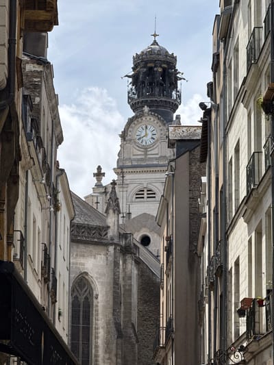 The image shows a street view in Nantes, France, with a prominent clock tower dominating the background. The clock tower is ornate, featuring a dome with statues of figures holding trumpets and a cross at its apex. A large clock face with Roman numerals is visible on the tower. To the left and right of the tower are narrow, aged buildings with windows and balconies. The architecture suggests a historic European city. The street itself is not clearly visible, but the perspective implies it is a pedestrian area or a narrow street between buildings. The sky is partly cloudy. The scene captures a daytime view of the urban landscape.