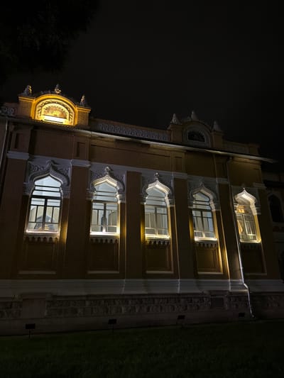 The image shows the Narzan Gallery in Kislovodsk, Russia, at night. The building is illuminated by artificial lights, highlighting its yellow brick facade and ornate architectural details. Several arched windows, framed with white decorative elements, are visible. Some windows are lit from within, casting a warm glow on the exterior. The top of the building features a decorative pediment with a backlit artwork. A dark tree branch extends from the upper left corner of the frame. The foreground consists of a dark lawn and some ground-level lights.