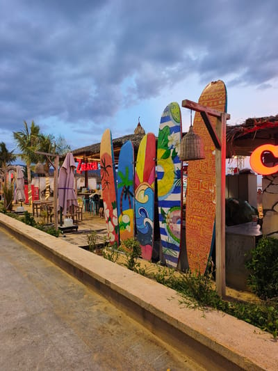 The image depicts a beachfront scene at dusk in An Nhơn, Vietnam, likely a restaurant or cafe. In the foreground, a concrete walkway runs alongside a raised planter box filled with green foliage. A row of vibrantly painted surfboards stands upright, serving as a decorative fence. Behind the surfboards, a thatched-roof structure, possibly a bar or seating area, is visible. Umbrellas are closed and stacked. A neon sign that reads "BEACH" in red letters is illuminated. The sky is overcast with dramatic clouds. The overall impression is of a relaxed, tropical atmosphere, transitioning from day to evening.