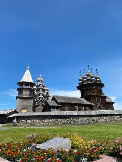 The image shows the Kizhi Pogost complex, a UNESCO World Heritage Site located on Kizhi Island in Karelia, Russia, which is near Boyarshchina. The complex features two wooden churches and a bell tower, all constructed in the traditional Russian architectural style. The larger church, the Church of the Transfiguration, is visible on the right with its distinctive onion domes, while the smaller Church of the Intercession and the bell tower are on the left. The buildings are made of wood and appear weathered, suggesting their age. They are surrounded by a low stone wall. In the foreground, there is a vibrant flower bed with orange and pink flowers, and a gravestone. Two individuals, a man and a woman, are walking on a path in the middle ground. The sky is a clear, bright blue with a few wispy clouds, and the sun appears to be shining, casting distinct shadows. The overall scene depicts an outdoor historical and cultural site under a pleasant weather.