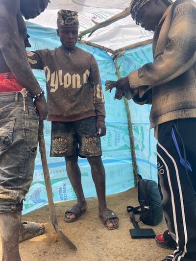 The image shows three individuals in a rudimentary shelter constructed from sticks and plastic sheeting. The shelter is erected over a surface that appears to be dirt or sand. One individual is lying down, holding a wooden stick; another stands over him. A third is slightly out of frame, reaching into the shelter. Their clothes are worn and dirty. Various objects are visible around the shelter: a small black bag, a black block, and an agricultural tool. Text on the bags used for the shelter includes the words "POWER HAM", "CBOMEK", "AGENE", and "DOWN AMERICA". The city mentioned in the request is not visible in the image.
