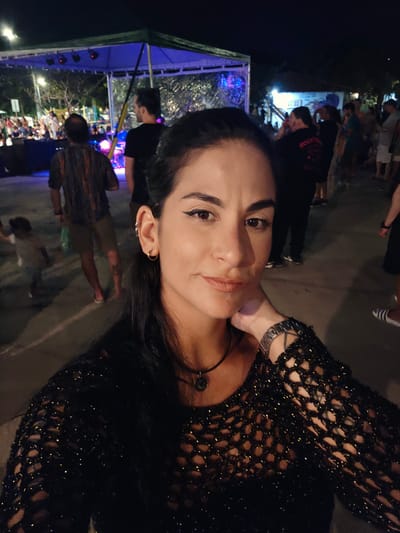 An eye-level selfie captures a woman in the foreground against the backdrop of a lively outdoor night event. The woman, with dark hair and a gentle smile, wears a black, shimmering, mesh-knit top and a necklace with a dark pendant. She has a metallic watch on her left wrist and multiple earrings on her right ear, including a helix cuff. Her gaze is directed towards the camera.
The background reveals a bustling scene at night, indicative of a public gathering, possibly a concert, festival, or market in Armação dos Búzios, Brazil. Numerous indistinct figures of people are visible, moving and congregating. To the right, a large blue tent or canopy structure is illuminated with bright blue stage lights. Other artificial lights dot the scene, suggesting a festive atmosphere. The ground appears to be a paved surface. One person in the background wears a shirt with the faint word "ROOTS" visible. The overall mood is casual and social.