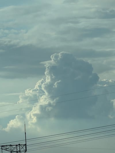 The image captures a cloudy sky over Tort-Kuduk, Kazakhstan. The clouds are predominantly white and gray, with some appearing darker and more dense. In the foreground, power lines stretch horizontally across the frame, with a metal tower structure visible at the bottom left. The overall scene is devoid of any people or specific activities, focusing solely on the atmospheric conditions and the man-made infrastructure.
