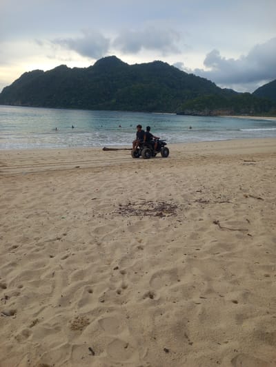 In Meunasahbale, Indonesia, a sandy beach is visible with the ocean in the background. Two individuals are seated on an all-terrain vehicle (ATV) parked on the sand. Several people are in the water, some appearing to be swimming. The water is a clear blue-green, with gentle waves approaching the shore. Lush green mountains rise in the distance, under a cloudy sky. The sand is textured with footprints and scattered debris, suggesting recent activity. The scene is captured from a low angle, emphasizing the expanse of the beach and the natural landscape.