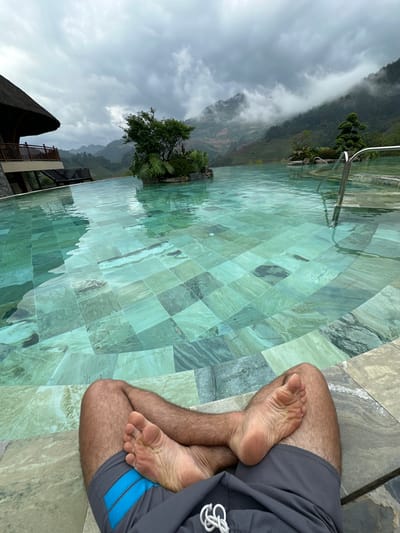The image captures a first-person perspective of a person relaxing by an infinity-edge swimming pool, overlooking a mountainous landscape in Púng Luông, Vietnam, under an overcast sky.
In the foreground, the bare legs and feet of a person are visible, resting on a tiled pool deck. The legs are tanned and hairy, crossed at the ankles. The person wears grey swimming shorts with two horizontal blue stripes on the left leg and a white drawstring on the right. The soles of the feet show visible wrinkles. The deck surface is composed of large, irregular grey and brown marble tiles.
The midground is dominated by a turquoise-green infinity pool with clear, rippled water. The pool bottom is tiled with a mosaic of square tiles in various shades of green, blue, and grey, exhibiting natural stone patterns. Two silver-colored pool ladders are partially submerged on the right side of the pool. Towards the center-left, a small landscaped island featuring green foliage and a small tree rises from the water.
The background presents a panoramic vista of lush green mountains, characteristic of Púng Luông's terrain. Patches of white fog or low clouds cling to the upper slopes of the mountains, particularly on the right side. Terraced fields are discernible on some mountain gradients. On the far left, a portion of a building with a dark, prominent thatched roof and wooden railings on a balcony is visible, suggesting resort architecture. The sky above is uniformly grey and overcast.