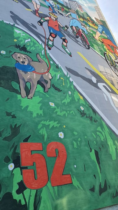 This is a detailed photograph of a vibrant mural painted on a wall, located in the city of Salihorsk, Belarus. The mural depicts a dynamic scene of outdoor recreation. In the foreground, a tan dog with a blue frisbee in its mouth is looking to the left, seemingly engaged with an unseen individual. The dog stands on a patch of green grass with white flowers. Behind the dog, a person on rollerblades, wearing a helmet, red shirt, and blue shorts, is being pulled by a red leash held by an unseen entity. Further in the background, several individuals are depicted engaging in various activities. One person is riding a red bicycle, another is on a yellow bicycle, and others are walking. The background also includes stylized buildings and a playground. The numbers "52" are prominently displayed in large, red, block letters in the lower left portion of the mural. The mural's perspective suggests it is painted on a vertical surface, possibly a building, and viewed from a slightly upward angle.