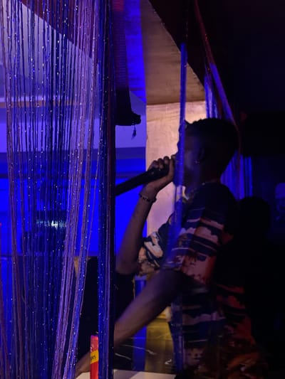 The image captures a dimly lit indoor scene, likely a performance venue in Durumi, Nigeria. A young Black male, positioned to the right of the frame and facing left, is holding a microphone to his mouth, suggesting he is performing or speaking. He is wearing a patterned shirt with blue, white, and orange elements. His right arm is raised, and his left hand is out of frame. To the left, a shimmering curtain of thin, dark strands, possibly tinsel or beaded string, hangs vertically, creating a layered effect and obscuring parts of the background. The environment is illuminated by vibrant blue and purple lights, casting a cool glow on the scene. In the background, other individuals are visible in silhouette, indicating an audience or other people present in the venue. The overall atmosphere appears energetic and engaging, characteristic of a live entertainment setting.
