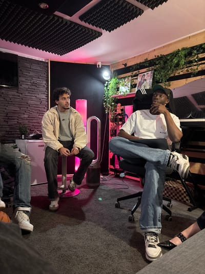 The scene depicts four individuals in what appears to be a music studio or creative space located in Boulogne-Billancourt, France. The interior features sound-dampening acoustic foam panels on the ceiling and upper left wall, a textured dark grey wall on the left, and horizontal light wood paneling adorned with artificial ivy on the right wall. The floor is covered with dark grey carpet.
In the left-center, a light-skinned male with dark, curly hair and a short beard is seated on a pink, circular-based stool. He wears a light-colored jacket over a grey Nike t-shirt, dark jeans, and light-colored sneakers. His hands are clasped in his lap, and he is looking intently towards the right, slightly upward. Behind him, a white mini-fridge or cabinet is visible.
To the right, a dark-skinned male is seated in a black office chair with wheels, facing left. He wears a black baseball cap backward, a white t-shirt with a small logo, blue jeans, and white and black Adidas sneakers. His legs are crossed, and he rests his chin on his right hand, looking left with a focused expression. Behind him, a black electronic keyboard is partially visible on a stand, and a wooden shelf displays a framed item resembling Michael Jackson's "Bad" album cover and a black Adidas cap.
Between these two primary figures, a tall, white and black Dyson air purifier stands on the floor. Ambient pink lighting emanates from a vertical light strip on the dark back wall and potentially from an unseen source on the right. A circular white floodlight is mounted on the right wall, and a small white security camera is affixed to the ceiling.
Two other individuals are partially visible: on the far left, the lower half of a person wearing ripped blue jeans and white high-top sneakers; and on the far right, the lower leg and foot of a person wearing dark trousers and black slip-on shoes with a strap. All visible individuals appear to be attentive.