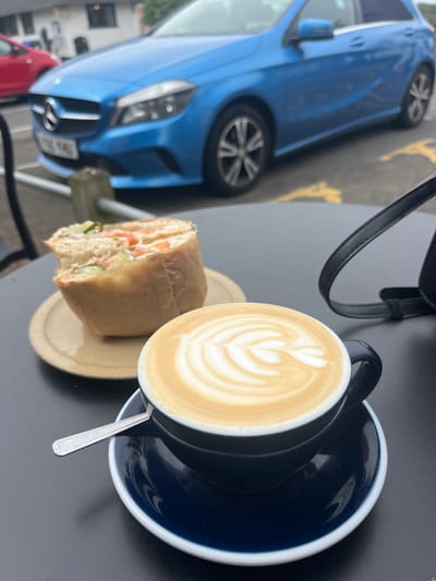 The image shows a close-up of a table setting with a latte in a dark blue cup and saucer, a sandwich partially wrapped in paper on a light-colored plate, and a spoon. A black bag strap is also visible. In the background, a blue car is parked diagonally across the frame, with a red car partially visible behind it. The location is indicated by the presence of the car's license plate as being in East Suffolk, United Kingdom.