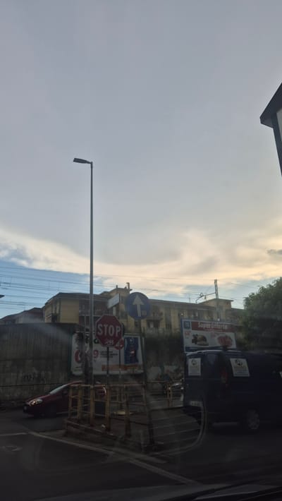 This image shows a street scene in Salerno, Italy, with a blue van and a red car visible. A tall street lamp stands prominently in the center of the frame, with a red "STOP" sign and a blue directional arrow sign beneath it. In the background, there are apartment buildings and railway tracks. The sky is overcast with some clouds. The van has two logos on its rear doors with the text "MANGO" and "SELEZIONATO". The red car is on the left side of the image, moving towards the intersection.