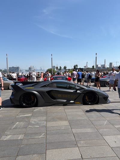 The image captures an outdoor car show on a sunny day, with a low-angle view of a matte grey Lamborghini Aventador as the primary subject in the foreground. The car features a prominent rear wing, black rims with yellow brake calipers, and yellow graphics on its side that read "VOSSEN" and "TOP X SECRET". In the background, a crowd of people are milling around, looking at various cars. The environment appears to be a paved area, possibly a waterfront promenade, with industrial elements visible in the distance, including cranes and buildings, suggesting an urban setting. The skyline and the overall atmosphere suggest the location is Gdańsk, Poland, likely near the port area. The sky is clear and blue with a faint contrail from an airplane.