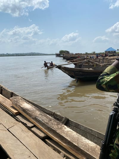 The image depicts a daytime scene at a riverfront in Gburuku, Nigeria, under a partially cloudy sky. In the immediate foreground, a wooden plank surface, possibly a dock or vessel deck, is visible, with several loose wooden planks resting upon it. The lower right portion of the frame shows the torso and arm of an individual dressed in green camouflage fatigues, holding a dark-colored rifle in a vertical position.
The mid-ground features a wide, calm body of muddy-brown water, identified as a river. Two individuals are observed within a small, single-canoe wooden boat on the river, with one individual actively paddling. Along the distant riverbank, several traditional wooden boats are moored. A group of individuals is scattered across the unpaved, sandy shoreline near these boats. Further right along the bank, a building with a light blue roof is visible. To the left of this building, a wooden pier or platform extends into the water. The distant horizon across the river shows a continuous line of dense green vegetation.