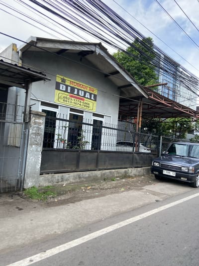 The image shows a house with a sign that reads "Rumah Ini Dijual" (This House is For Sale) in the Kecamatan Margahayu, Indonesia. The sign also contains two phone numbers. The house is made of concrete and has a black metal fence in front of it. There is a black car parked on the side of the road in front of the house. The car has a license plate that reads "D 1088 CZ". The street is paved and has a white line painted on the edge. There are several power lines overhead.  The house appears to be located on a residential street.  The environment is cloudy, and the sky is overcast.