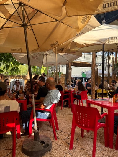 This image depicts a bustling outdoor dining area, likely a restaurant or bar patio, in San Pedro Garza García, Mexico, under bright daylight. Numerous people, primarily adults, are seated at red plastic tables and chairs, engaging in eating, drinking, and socializing. The scene is well-shaded by several large, beige canvas umbrellas.
On the tables, a variety of drinks, bottles, and glasses are visible, suggesting a casual meal or refreshment. The flooring is a light-colored, textured surface, possibly gravel or stone paving. In the background, beyond the immediate dining area, wooden support beams frame the space, alongside greenery from trees, and utility lines. A white truck is partially visible further in the background. Overhead, string lights are installed, hinting at evening use, and a fan is mounted on a wooden post. A heavy black base anchors one of the umbrella poles, and a green sign clearly reads "SALIDA" (Exit).
The time of day appears to be daytime, likely afternoon, given the bright sunlight and the essential function of the shade umbrellas. The weather seems clear and sunny. People are dressed in casual attire and appear relaxed, contributing to a convivial atmosphere. Visible text on the large umbrellas includes "PACIFICO" and "STELLA ARTOIS," both well-known beer brands, along with partial words like "CILICO" and "TEICO."