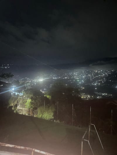 A dark, night view reveals a cityscape of numerous bright lights sprawling across the landscape. The lights could be from buildings or vehicles. To the right, trees and a metal structure are vaguely visible in the dimness. A cloud-filled, dark sky forms the backdrop. The perspective gives the impression of an elevated vantage point overlooking the luminous urban scene.