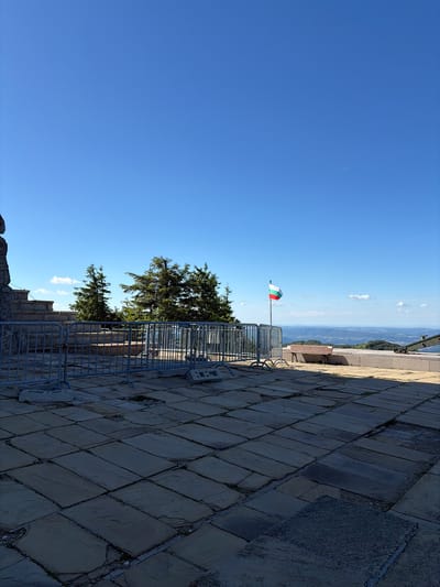 This image captures a sunny day at the Monument to the Freedom of Bulgaria, also known as the Shipka Monument, located in the city of Shipka, Bulgaria. The foreground features a stone-tiled plaza, with some tiles cracked or missing, suggesting wear and tear. A temporary metal barrier fence is set up, with concrete blocks used as stabilizers. Behind the fence, a stone monument or structure is visible on the left, with tiered steps leading up.
In the middle ground, several tall coniferous trees stand against a clear blue sky, with a few scattered white clouds. A Bulgarian flag, featuring white, green, and red horizontal stripes, is prominently displayed on a flagpole, waving gently. Further back, a wooden bench is placed on a slightly elevated platform. The background reveals a vast, rolling landscape of hills and valleys, suggesting a mountainous or elevated location. The overall impression is one of a scenic overlook and a historical monument.