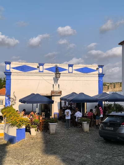 The image depicts an outdoor dining area in the historic city of Óbidos, Portugal. The scene is set on a cobblestone street in front of a building with white walls and blue architectural accents, featuring a diamond pattern above the windows. A sign indicates it is "1 DE DEZEMBRO - CAFÉ - RESTAURANTE". Several dark blue umbrellas provide shade over tables where groups of people are seated, appearing to be dining and socializing. The people are diverse in age and gender, casually dressed. A black Nissan Leaf electric car is parked on the right side of the frame. Lush yellow flowers are visible in a planter to the left, adding color to the scene. In the background, sections of the ancient castle walls of Óbidos are visible on a hill, suggesting the restaurant is located within or near the fortified town. The sky above is a mix of blue with scattered white clouds, and the lighting suggests it is late afternoon.