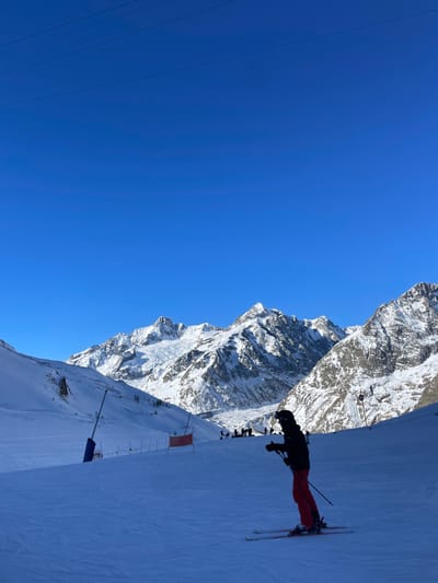 The image displays a bright, sunny day on a snow-covered mountain slope in Courmayeur, Italy. In the foreground, positioned on the right, a skier is visible, dressed in a black jacket, red pants, and a helmet, standing with skis and poles on the groomed snow. The vast majority of the left side of the frame is filled with a clear, deep blue sky, indicating excellent weather.

In the background, rugged, snow-capped mountains rise majestically, showcasing their rocky and snowy peaks and slopes under the bright sunlight. Along the ski slope, particularly further down, other smaller figures of people or skiers are discernible, along with what appears to be orange safety netting or flags. Thin lines, likely ski lift cables or power lines, cut across the upper left section of the sky. The scene depicts a typical winter sports activity with perfect visibility and conditions, likely during mid-day or early afternoon given the strong sunlight and clear shadows. No legible text is visible in the image.