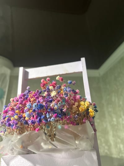 The scene depicts an indoor environment in Dmitriyevskaya, Russia. The primary subject is a dense arrangement of small, dried, multi-colored flowers, displaying hues of pink, blue, purple, yellow, and white. These flowers are contained within an open, light-colored wooden frame, which appears to be lavender or white. Their stems are gathered and secured at the base by translucent white wrapping material inside the frame. In the background to the right, a textured light green wall is visible, topped by a white crown molding. The upper section of the image shows a dark gray ceiling, separated from the wall by a distinct white trim line. An out-of-focus, light-colored object, possibly blue or teal, is partially visible in the far left background. The scene is illuminated by artificial light.