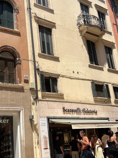 The image shows a street view of a building in Verona, Italy. The building has a light yellow facade with several windows, some of which have dark green shutters. A balcony with an ornate wrought-iron railing juts out from the second story. Below, a sign reads "Scaravelli Gelateria" and "Caffetteria e Pasticceria". Several people are visible in front of the gelateria, some entering and others browsing outside. The ground floor also features shop windows, one with "ker" visible on its signage, suggesting a retail establishment. A street number "46" is displayed on the building's facade. The overall scene depicts a bustling street in a European city.