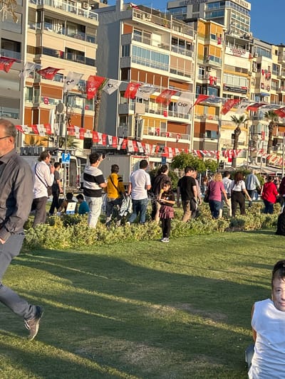 The image captures a street scene in İzmir, Türkiye, specifically at the coordinates 38.438 latitude, 27.141 longitude. It shows a group of people standing on a grassy area, likely a park or a promenade, adjacent to a street lined with multi-story apartment buildings. The buildings are adorned with numerous flags, primarily red with white lettering, and some white and red flags, possibly political or celebratory in nature. The people, diverse in age and attire, appear to be engaged in conversation, perhaps observing something beyond the frame. There are also a few people seated on the grass. The overall scene depicts a typical day in the city, with people going about their daily lives. The buildings, flags, and greenery suggest a vibrant and bustling urban setting.