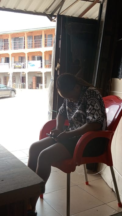 The image presents a daytime scene in Ogbunike, Nigeria, viewed from a shaded interior space looking out onto a brightly lit street. In the immediate foreground, a person with braided hair, wearing a patterned dark and light short-sleeved top and dark shorts, is seated in a red plastic chair. The individual is looking down and actively engaged with a black smartphone held in both hands. A dark wooden table corner is visible on the lower left. The floor beneath the chair is tiled with light-colored square tiles. Overhead, a corrugated metal roof structure is partially visible, and on the right, dark vertical objects, possibly door frames or shelving, are present.
The outdoor midground reveals a multi-story building, likely residential or mixed-use, with an orange-colored upper facade featuring multiple balconies with metal railings. The ground floor is a lighter grey. Numerous open and closed entryways are visible on the building, some appearing to display various doors. Text is visible on signs, including "Cee Tech" on the ground floor left and another partial banner. A dark grey or silver sedan is parked on the left side of the outdoor area, and scaffolding or ladders are positioned near the building's ground level.