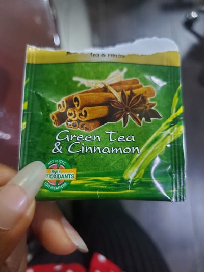 A hand with long, light-colored fingernails is holding a green tea bag packet. The packet has an image of cinnamon sticks and star anise on it, and text that reads "Green Tea & Cinnamon." There is also text indicating it is "High in TIOXIDANTS" and "LOW IN CAFFEINE." The background is out of focus, but appears to be an indoor setting. The image is not specific to Gwagwada, Nigeria.