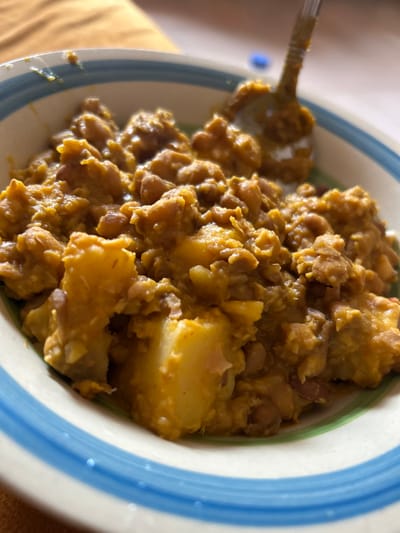 A close-up captures a bowl filled with a hearty stew. Brown beans and chunks of yellow potatoes are visible. A metal spoon rests in the bowl, partially covered in the stew's thick sauce. The bowl has a white interior and rim, decorated with green and blue stripes. The background is a soft, warm yellow. The stew has a thick and creamy texture.