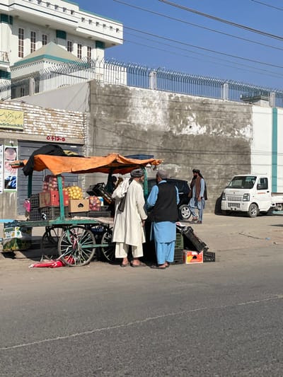 Three men stand beside a fruit cart laden with apples, oranges, and various crates. One man wears a light blue shalwar kameez with a dark vest, while another is clad in a white shalwar kameez. A third man stands partially obscured behind the cart. The cart has an orange tarp offering shade. A white van is parked nearby along a building, and the scene is set on a sunny street in a town.