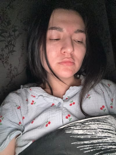 The image shows a female individual indoors, likely in Kurovskoye, Russia, resting in a dimly lit setting. Her head is tilted slightly to her right, and her eyes are closed, indicating sleep or repose. Her lips are slightly parted. She has dark hair, which is loose and falls around her face and shoulders. The individual is wearing a light-colored, possibly pale blue or grey, textured button-down shirt with a ruffled collar and a repeated pattern of small red cherries with green stems. A dark pillow or blanket with a white abstract pattern is visible in the lower right portion of the frame, partially covering her torso. The background on the left side reveals a wall adorned with a subtle, dark floral or botanical pattern, consistent with wallpaper. No other individuals or active interactions are visible.
