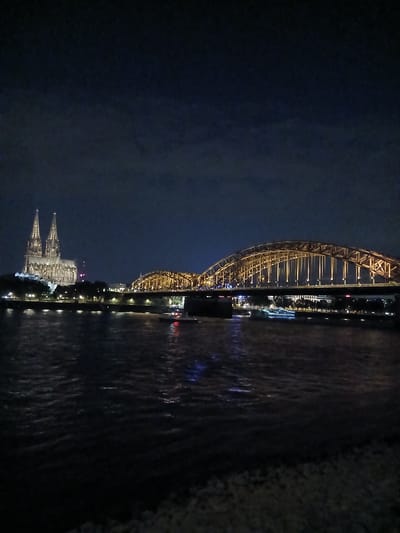 The image shows a nighttime view of the Hohenzollern Bridge and Cologne Cathedral in Cologne, Germany, as seen from across the Rhine River. The cathedral, with its twin spires, is illuminated and stands to the left of the frame. The Hohenzollern Bridge, a railway bridge known for its arches and decorative elements, is lit with golden lights and spans the river in the middle and right of the frame. The river itself reflects the lights of the city and the bridge, creating a shimmering effect on the dark water. Several boats are visible on the river, some with their own lights. The sky is dark with some faint clouds. The overall scene depicts a well-known landmark at night.