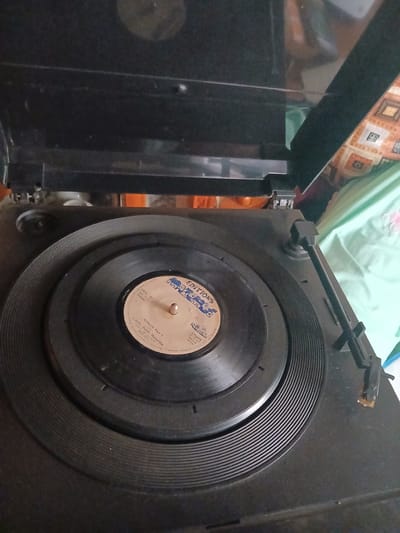 The image shows a close-up, overhead view of a black record player with a vinyl record on its turntable. The record has a light-colored label with blue text. The label indicates "EDITIONS", "IMA 31", "Made in A Stereo", "NANGA Part 1", "Lorch Super Mazembe", and "Lingala All". The record player is positioned on a surface that appears to be indoors, possibly in a room with a patterned textile visible in the background. There are no people or visible action in the scene. The environment appears to be a domestic setting. The location is described as Witeithie ward, Kenya.