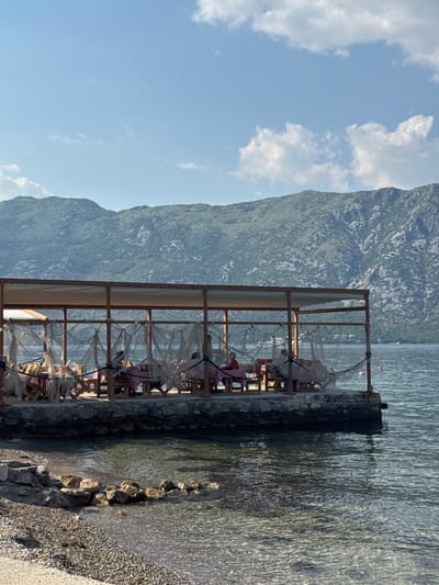 The image captures a waterside restaurant in Prčanj, Montenegro. The restaurant is situated on a stone embankment extending into the bay. Its structure consists of a simple wooden frame supporting a roof, with seating arranged inside. Nets or light fabric hang around the edges. To the left, a mountain rises, partially obscured by a sky with scattered clouds. The water laps at the embankment's edge. Rocks and gravel form the shoreline. The image is vertically oriented.