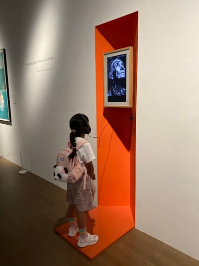 A young East Asian girl with a black braided ponytail, wearing headphones, a white t-shirt, pink shorts, pink and white striped socks, and pink sneakers, stands facing an orange wall in a museum. She has a light pink backpack with a black and white panda plush attached. On the orange wall, a digital frame displays a black and white portrait of an elderly person looking upwards. A quote in Chinese text is visible on the white wall to the left of the digital frame, with an English translation below it. The floor is made of dark wood. The scene appears to be in a museum located in Jinniu District, China.