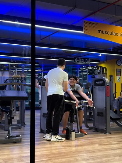 The image depicts an indoor gym environment with bright blue and yellow lighting. In the foreground, two young men are visible. One is standing and wearing a white t-shirt and black pants, while the other is seated on a weight machine, looking towards the standing man. The seated man is wearing a grey t-shirt and shorts and has a slight smile. The gym is equipped with various weight machines, one of which is labeled "HAMMER STRENGTH." The floor is made of light-colored wood. In the background, a sign with the word "SALIDA" (exit in Spanish) and an arrow pointing left is visible, as well as another sign that appears to say "musculación" (weight training in Spanish). The overall atmosphere suggests a typical gym setting. This scene is likely located in the city of San Isidro, Peru, a district known for its modern commercial and residential buildings, which often house fitness centers.