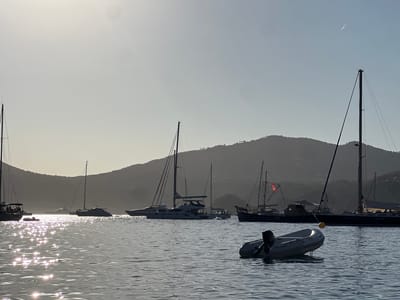 The image shows a harbor scene at a low sun angle, likely during sunset or sunrise, with the sun creating a shimmering effect on the water. Several sailboats and yachts are anchored in the water, with their masts silhouetted against a hazy sky. In the foreground, a small inflatable dinghy with an outboard motor is floating. In the background, a hilly coastline is visible, also silhouetted. The scene appears to be in a bay or sheltered cove. The location is likely Sant Josep de sa Talaia, Spain, which is known for its scenic coastline and harbors. There are no people clearly visible in the image. The overall atmosphere is serene and peaceful, with the natural light creating a soft, diffused illumination.