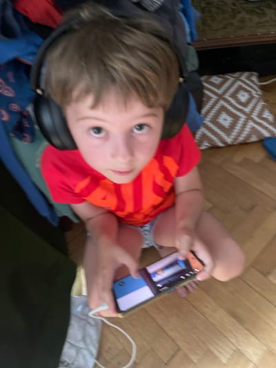 A young boy wearing headphones and a red t-shirt is sitting on a wooden floor, holding a smartphone. The boy is looking at the phone, which appears to be a game or video. The background is cluttered with clothes and a patterned pillow. The lighting suggests it is daytime. The image does not contain enough information to determine the location within the city of Bydgoszcz, Poland.