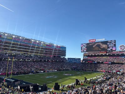 Levi's Stadium hosts Super Bowl 50, filled with spectators. The stadium's towering structure is adorned with "SUPER BOWL L" signage. A large video screen displays an image. The football field is marked with "SEATTLE" in one end zone. A jet stream cuts across a clear blue sky. A plane travels leaving behind white smoke.