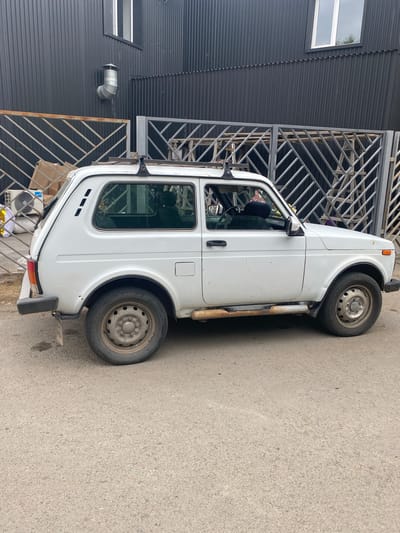 A white Lada Niva is parked on an asphalt road in front of a dark corrugated metal building with a fence. The car is facing right and shows signs of wear, with rust on the side rocker panels and some dirt on the body. The building has at least two windows and a silver ventilation pipe visible on its exterior. The fence is made of grey metal with a diamond pattern. In the background, to the left of the car, there is an air conditioning unit and some cardboard boxes. The scene appears to be an outdoor area in the city of Ekibastuz, Kazakhstan. There are no people clearly visible in the image.
