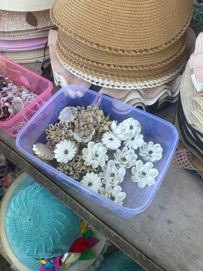 In Bira, Indonesia, on a weathered wooden surface, a stall displays various handmade crafts. Stacked straw hats in shades of beige and white dominate the upper portion of the image. In the foreground, a purple plastic container is filled with decorative items made from seashells. These include several flower-shaped ornaments and clusters of small shells arranged in circular patterns. To the left of the purple container, a pink plastic container holds a collection of colorful beads and small charms, possibly part of jewelry-making supplies. Below the containers, a light blue crocheted hat with a woven brim and decorative fabric flowers is visible. The overall scene suggests a market or craft stall offering beach-themed accessories.
