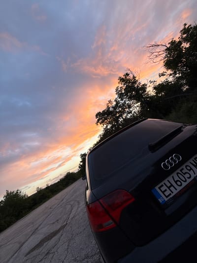 The image displays the rear section of a black Audi car parked outdoors, set against a dramatic sunset or sunrise sky. The photograph is oriented with the horizon line diagonally across the frame.

The main subject is a dark-colored Audi vehicle, with its distinctive four-ring emblem clearly visible on the trunk lid. The car's right taillight, a prominent red rectangle, is visible towards the bottom right corner. A Bulgarian license plate is mounted on the rear, showing the country code "BG" and the alphanumeric sequence "EH 6059". Below the main characters, the text "КРЪВНА ГРУПА" (Blood Group) is also legible on the plate, likely a common sticker in the region.

The background is dominated by a vibrant sky, painted with hues of soft purples, blues, and an intense band of oranges, pinks, and yellows where the sun's low light illuminates scattered clouds. Dark silhouettes of trees and foliage frame the sky from the top and left, suggesting a natural, possibly wooded, environment. The ground beneath the car and extending to the lower right appears to be an asphalt surface, possibly a road or parking area, with subtle tire marks visible.

The scene suggests a peaceful moment at either dawn or dusk, characterized by the warm, natural lighting. There are no people visible, and no specific activity is taking place; the car appears to be stationary. The location cues from the license plate confirm the setting in Bulgaria, specifically in or around Tuchenitsa as provided.