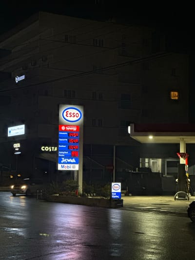 The image captures a nighttime view of an Esso gas station and its surroundings in Egkomi, Nicosia, Cyprus. The scene is dominated by a brightly lit Esso sign displaying fuel prices against the dark backdrop of a multi-story building. The ground, a wet asphalt road, reflects the artificial lights, suggesting recent rainfall.

The prominent gas station sign features the circular Esso logo at the top. Below it, listed prices are: "Synergy" at 1.459, another fuel type at 1.549, a third at 1.549, and "Jet Wash" at 1.599, followed by "Mobil II". A "COSTA" sign is also visible beneath the price display. To the left, another illuminated sign reads "Payams" and below it, partially visible, a sign reads "Prams & Mermaids". In the middle distance on the right, a smaller, illuminated Esso sign indicates "Exit Έξοδος" (Exit). The large building in the background has several windows, with one on an upper floor being lit. No people or active vehicles are clearly visible, contributing to a quiet, late-night atmosphere.