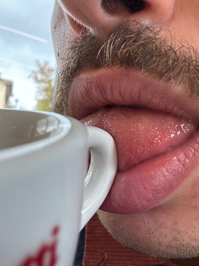 A close-up, angled photograph depicts the lower facial area of an individual. A dense, light-to-medium brown mustache covers the region above the upper lip. The pink lips are partially open, revealing an extended, moist, pinkish tongue with visible papillae, which is pressed against the bright white ceramic handle of a mug. The upper lip exhibits fine vertical lines. Light stubble is visible on the chin and adjacent facial skin. A blurred red logo or text is partially visible on the side of the white mug. The out-of-focus background features a light grey sky, indistinct light-colored architecture, and blurry autumnal foliage with yellow and orange tones, suggesting an outdoor environment or a view through a window. A segment of reddish-brown fabric, consistent with clothing, is present at the bottom of the frame. The scene is located in Anzola dell'Emilia, Italy.