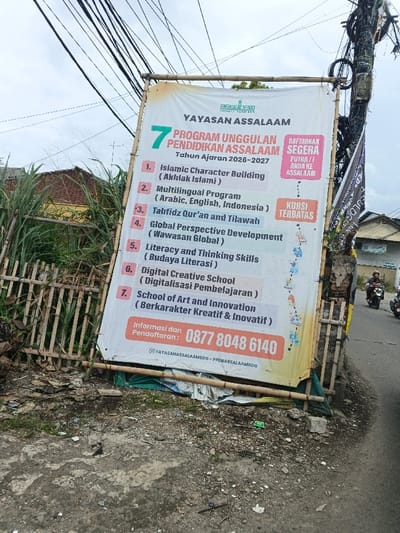 This image depicts a roadside scene in Margahayu, Indonesia, under an overcast sky. A large, rectangular white fabric banner with yellowed edges, supported by bamboo poles, stands prominently on the left side of the frame. The banner advertises "YAYASAN ASSALAAM" and its "7 PROGRAM UNGGULAN PENDIDIKAN ASSALAAM" for the academic year 2026-2027, listing programs such as Islamic Character Building, a Multilingual Program (Arabic, English, Indonesian), Tahfidz Qur'an and Tilawah, Global Perspective Development, Literacy and Thinking Skills, Digital Creative School, and School of Art and Innovation. It displays calls to action including "DAFTARKAN SEGERA PUTRA/I ANDA KE ASSALAAM" and "KURSI TERBATAS," along with a contact number (0877 8048 6140) and social media handles.
To the left of the banner, a bamboo fence runs horizontally, partially obscuring tall, dense green reedy plants. Behind these plants, a yellowish-brown building with a red-tiled roof is visible. The immediate foreground consists of uneven dirt and concrete ground scattered with minor debris. To the right of the banner, a utility pole heavily laden with numerous black cables and an additional dark, partially visible banner is present. A paved asphalt road extends from the mid-ground towards the background on the right side of the image. Two individuals are observed riding separate motorbikes on this road, moving away from the viewer. Both individuals are wearing helmets. In the further background on the right, another light-colored building is visible. Numerous black utility wires crisscross the upper portion of the frame against the cloudy sky.